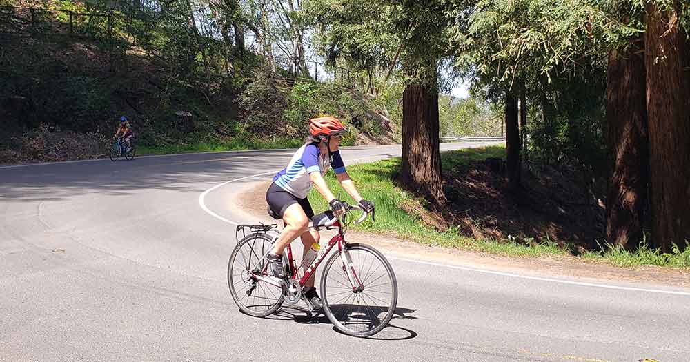 Photo of a woman riding her road bike on a country road in the hills with burn trees on the hillsides around her.